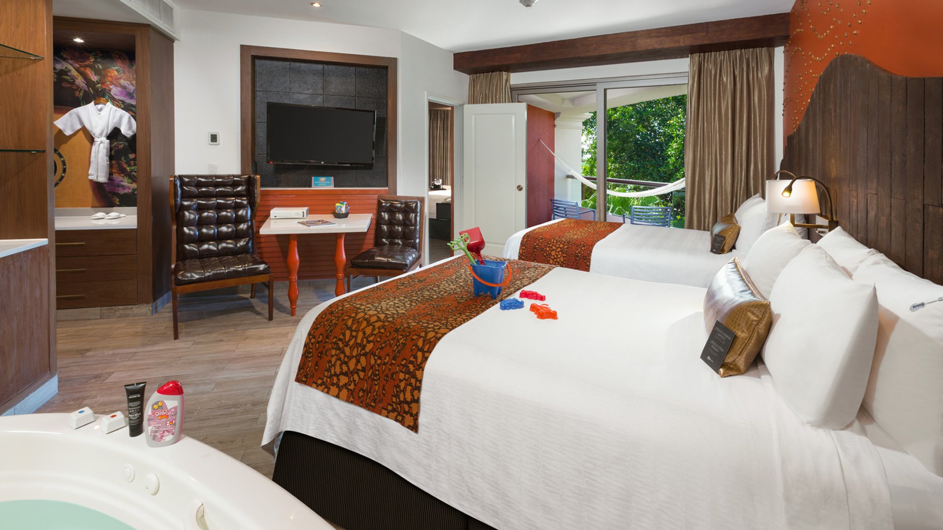 Spacious hotel room with two beds, TV, indoor hot tub, toys on a bed, and a patio with a hammock and garden view.